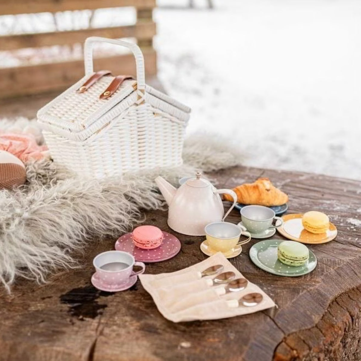 A styled picnic scene outdoors on a rustic wooden table with a white wicker picnic basket, a pink teapot, pastel cups and saucers, macarons, a croissant, and a fur throw blanket, with a snowy background.