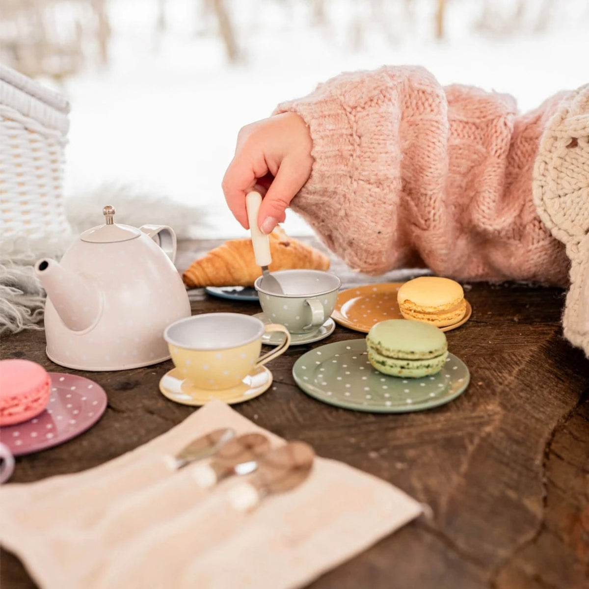 A cozy outdoor tea-time setup on a rustic wooden table with pastel-colored teacups, a teapot, macarons, and a child in a pink knit sweater stirring with a spoon. A white wicker basket and a blurred snowy background complete the scene.