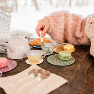 A cozy outdoor tea-time setup on a rustic wooden table with pastel-colored teacups, a teapot, macarons, and a child in a pink knit sweater stirring with a spoon. A white wicker basket and a blurred snowy background complete the scene.