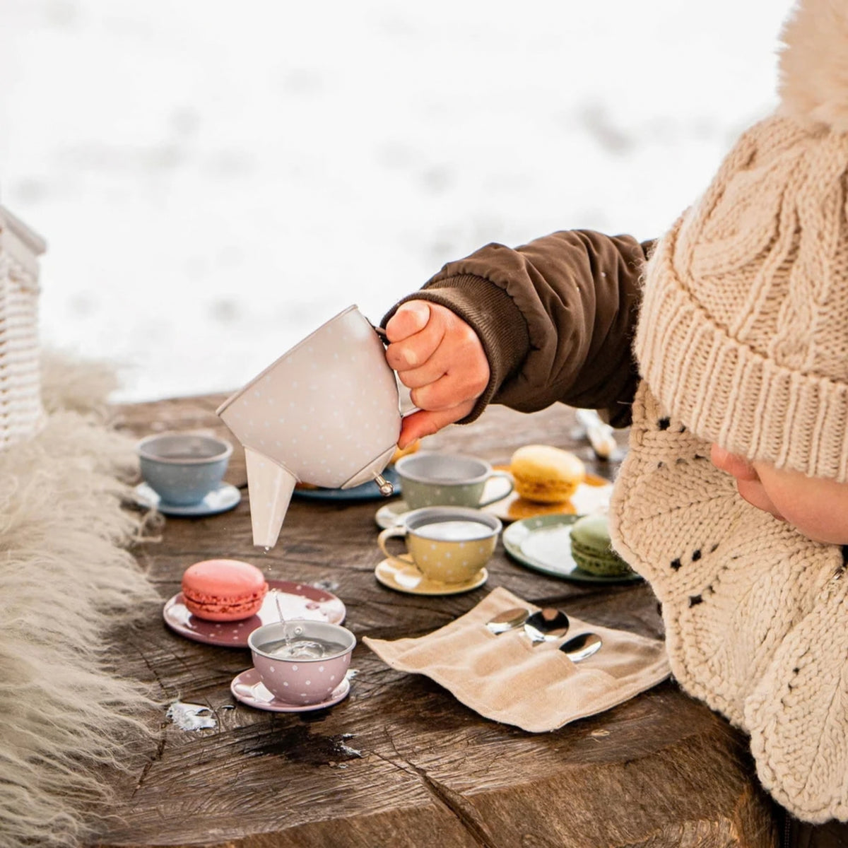 A child wearing a cream knit beanie and brown jacket pours hot water from a teapot into a pink teacup on a rustic wooden table, set up with macarons and other teacups in an outdoor snowy setting.