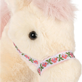 Close-up of a plush toy horse with cream-colored fur, a pink mane, and a pink floral bridle with green leaves and pink flowers.