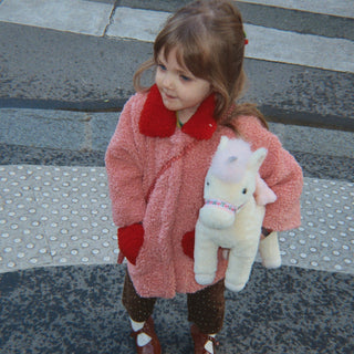 A young child wearing a fuzzy pink coat with red trim and holding a white plush unicorn, standing on a city crosswalk.