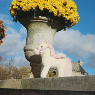 A plush white and pink unicorn toy with a pink patterned halter stands outdoors next to a large stone planter filled with yellow flowers under a blue sky.
