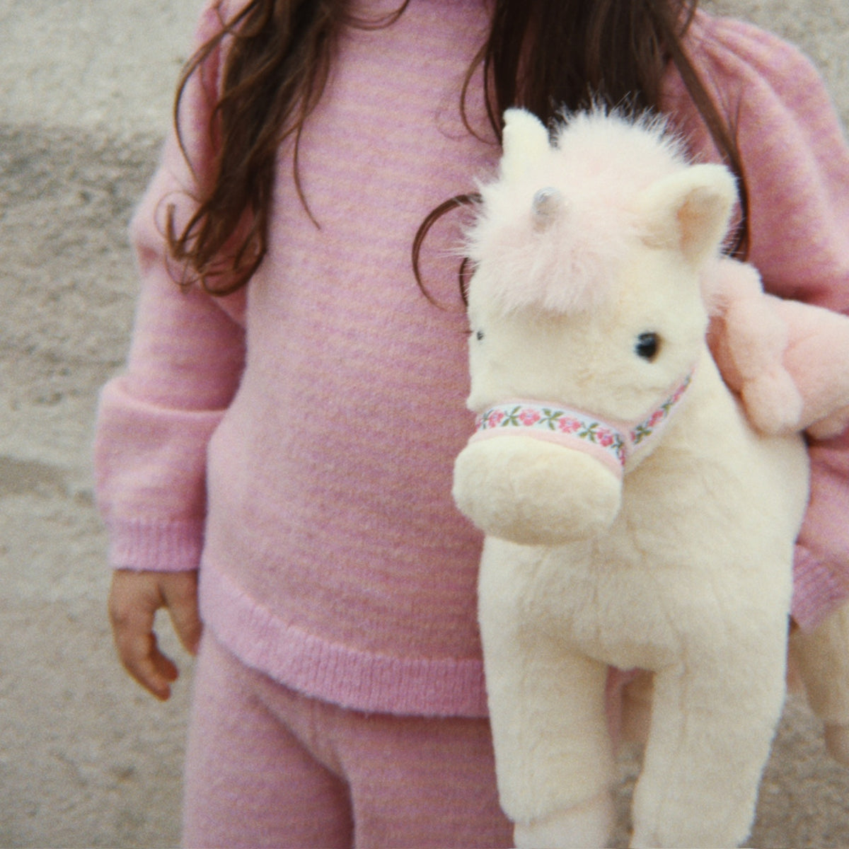 A child wearing a soft pink knit sweater and matching pants holds a fluffy cream-colored plush unicorn toy with a floral-patterned halter.
