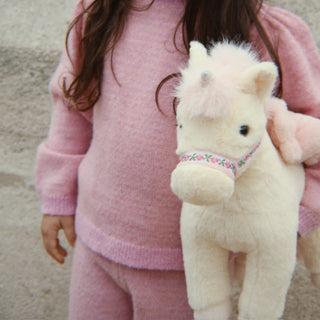 A child wearing a soft pink knit sweater and matching pants holds a fluffy cream-colored plush unicorn toy with a floral-patterned halter.