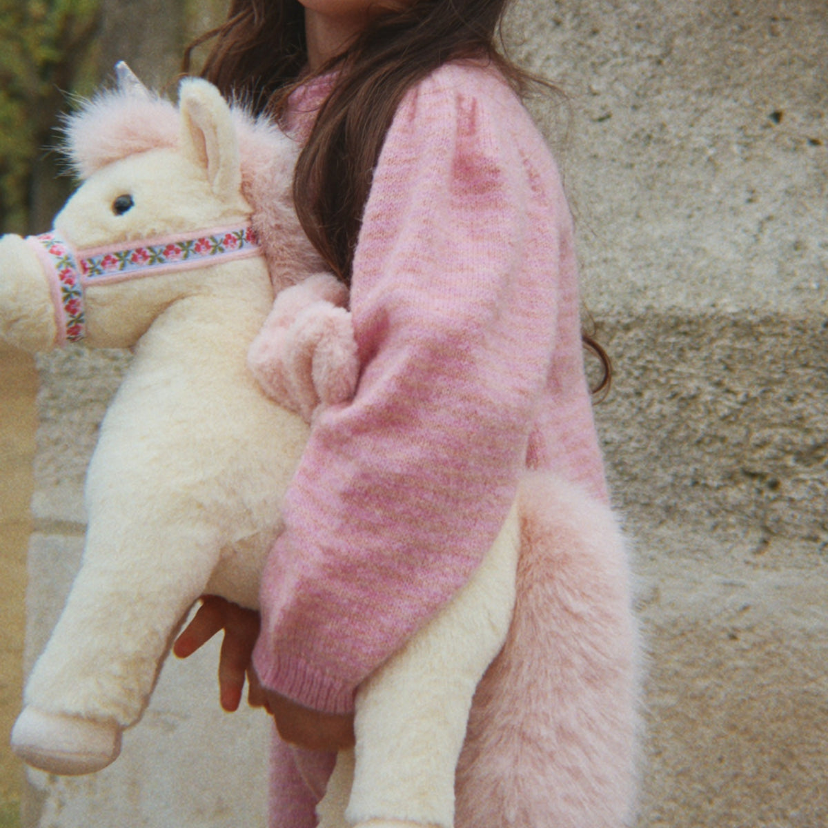 A child wearing a fuzzy pink sweater holds a plush cream-colored horse toy with a patterned bridle against a stone wall background.
