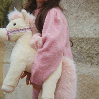 A child wearing a fuzzy pink sweater holds a plush cream-colored horse toy with a patterned bridle against a stone wall background.