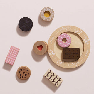 A set of wooden toy biscuits arranged on a pale pink background, including a round tray with a pink iced ring and a brown Bourbon chocolate biscuit, plus separate cookies, and a pink wafer.