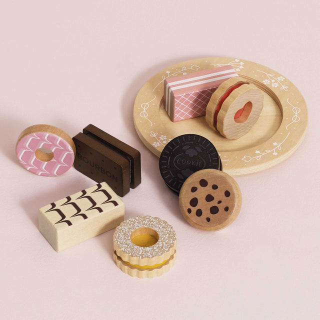 A set of wooden toy biscuits arranged on a round beige plate with floral detailing, including a pink iced ring, a dark chocolate cookie, a patterned rectangular piece, a heart-shaped cookie, and a brown rectangular piece labeled BOURBON, photographed on a pink background.
