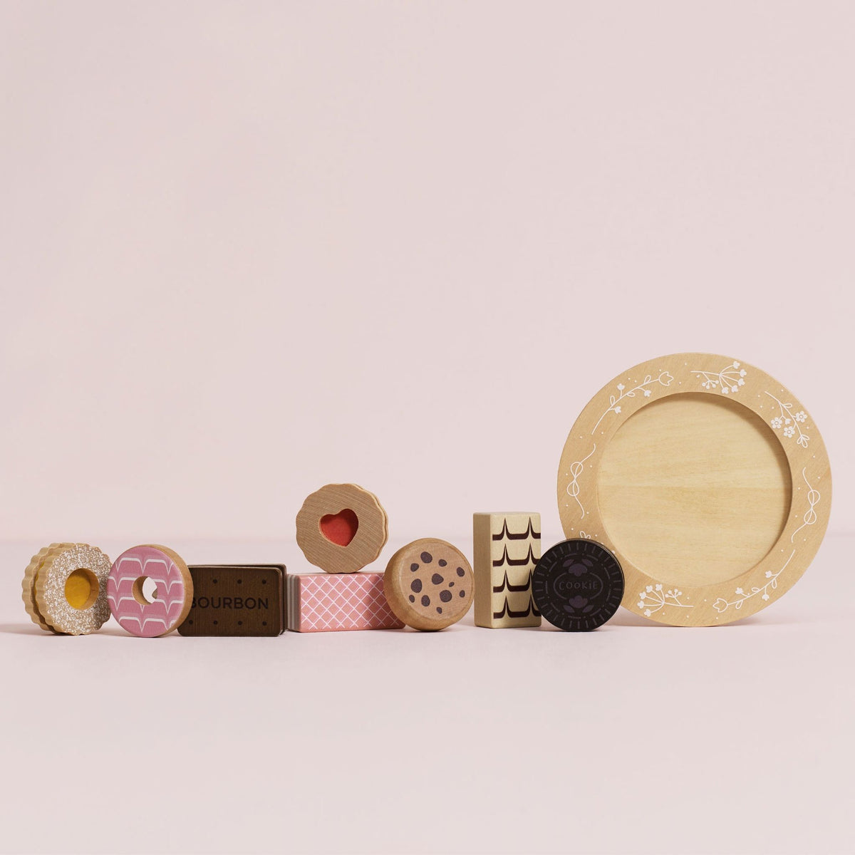 A set of wooden toy biscuits arranged on a round beige plate with floral detailing, including a pink iced ring, a dark chocolate cookie, a patterned rectangular piece, a heart-shaped cookie, and a brown rectangular piece labeled BOURBON, photographed on a pink background.