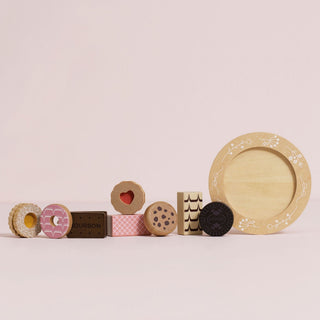 A set of wooden toy biscuits arranged on a round beige plate with floral detailing, including a pink iced ring, a dark chocolate cookie, a patterned rectangular piece, a heart-shaped cookie, and a brown rectangular piece labeled BOURBON, photographed on a pink background.