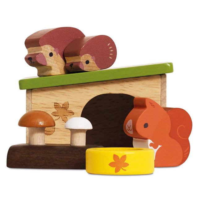 A wooden toy play set featuring a small shelter with a green roof, two hedghog figures on top and one red squirrel beside it, two mushrooms on a dark base, and a yellow water bowl with a flower, all photographed on a white background.