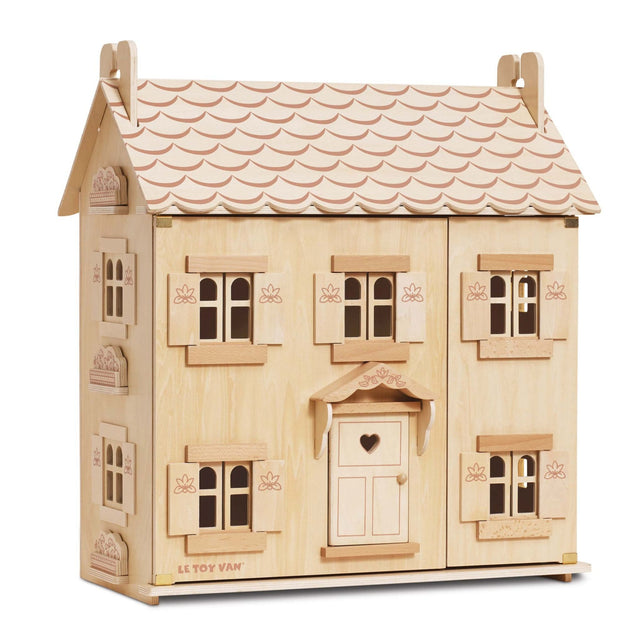 A wooden three-storey dollhouse with a light natural wood finish, featuring a scalloped light pink roof, multiple windows with shutters, and a central door with a heart-shaped window.