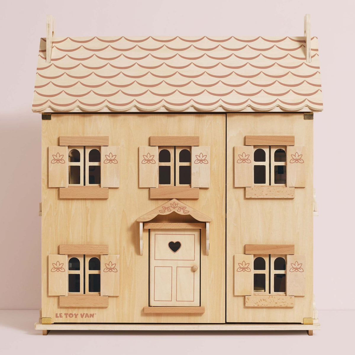 A wooden three-storey dollhouse with a light natural wood finish, featuring a scalloped light pink roof, multiple windows with shutters, and a central door with a heart-shaped window.