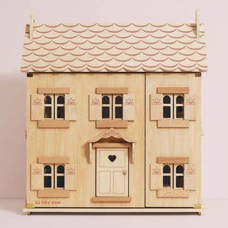 A wooden three-storey dollhouse with a light natural wood finish, featuring a scalloped light pink roof, multiple windows with shutters, and a central door with a heart-shaped window.