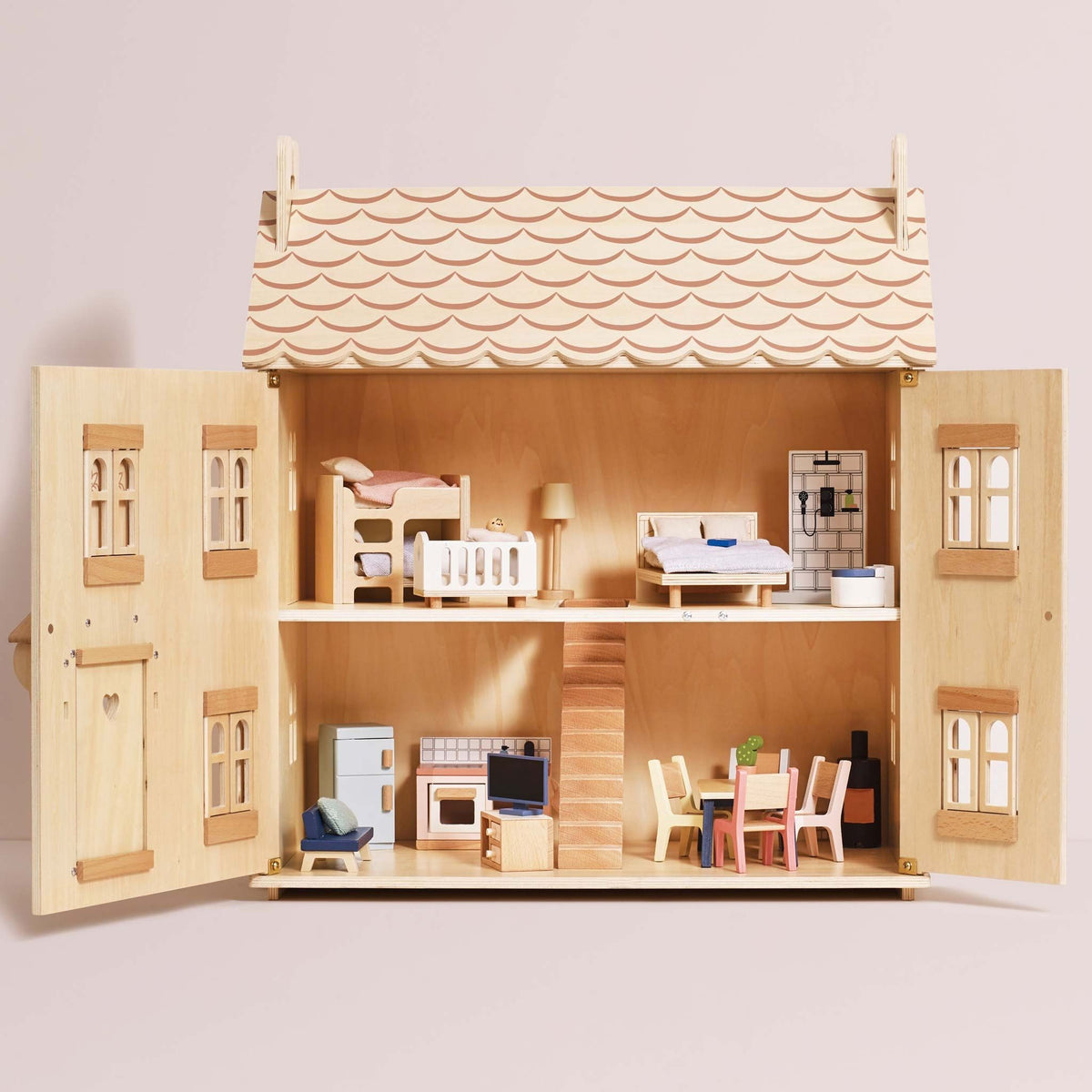 A wooden dollhouse with open doors, showcasing a two-story interior with furniture pieces, beds, a kitchen, and a staircase, set against a solid pale pink background.
