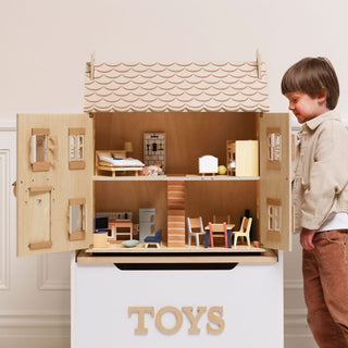 A young boy stands beside a large wooden toy house with open doors, revealing detailed miniature rooms, furniture, and a painted tile roof, set against a light beige wall with a white storage box labeled 'TOYS' below.