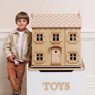 A young boy stands next to a large wooden dollhouse toy on a white display stand. The house has a light natural wood finish, a light pink painted scalloped roof, and a small heart-shaped cutout on the front door.