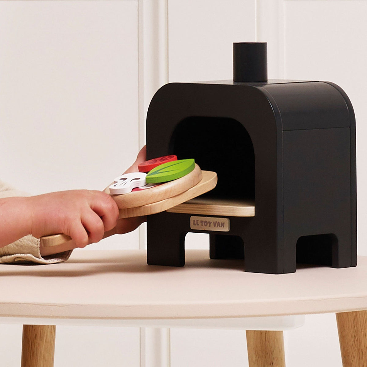 Black toy wooden oven with a child's hand holding a wooden peel with a toy pizza.