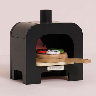 A toy wooden pizza oven by Le Toy Van, featuring a black oven body with a wooden peel and a small window showing colorful toy pizza , on a light pink background.