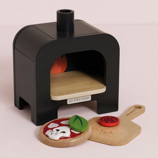 A children's wooden toy pizza oven set on a pale pink background, featuring a black oven body, light wood interior shelf, small orange flame detail, toy pizza, and a wooden pizza peel. The brand 'LE TOY VAN' is visible on the oven front.