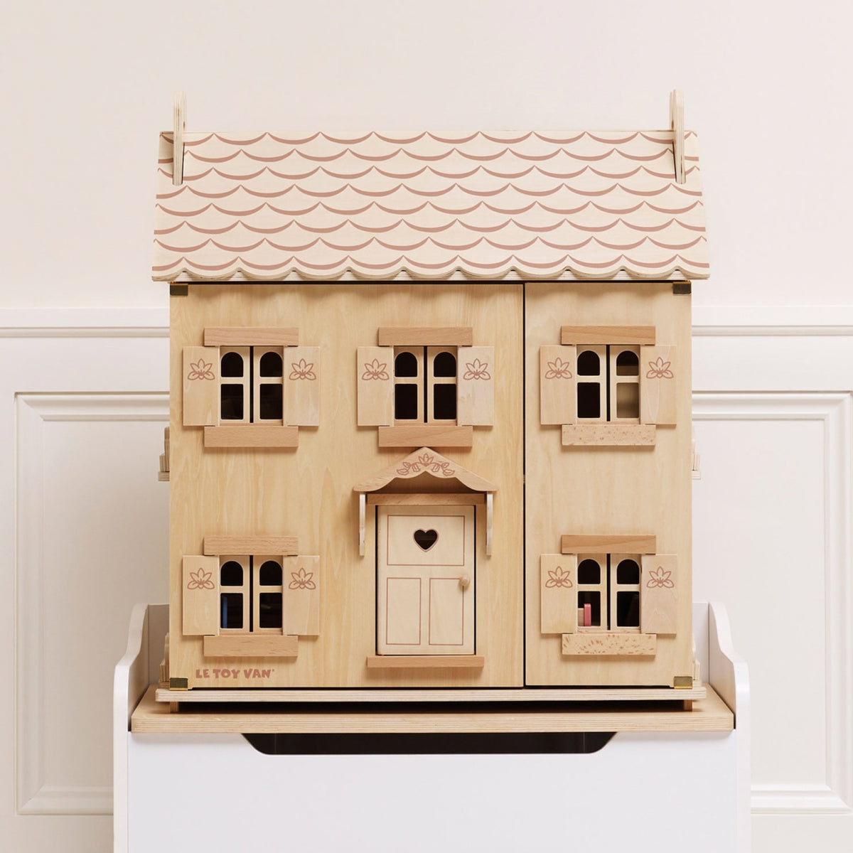 A wooden dollhouse with a scalloped roof, multiple windows, shutters, and a central door with a heart hole, displayed on a white stand against a white paneled wall.