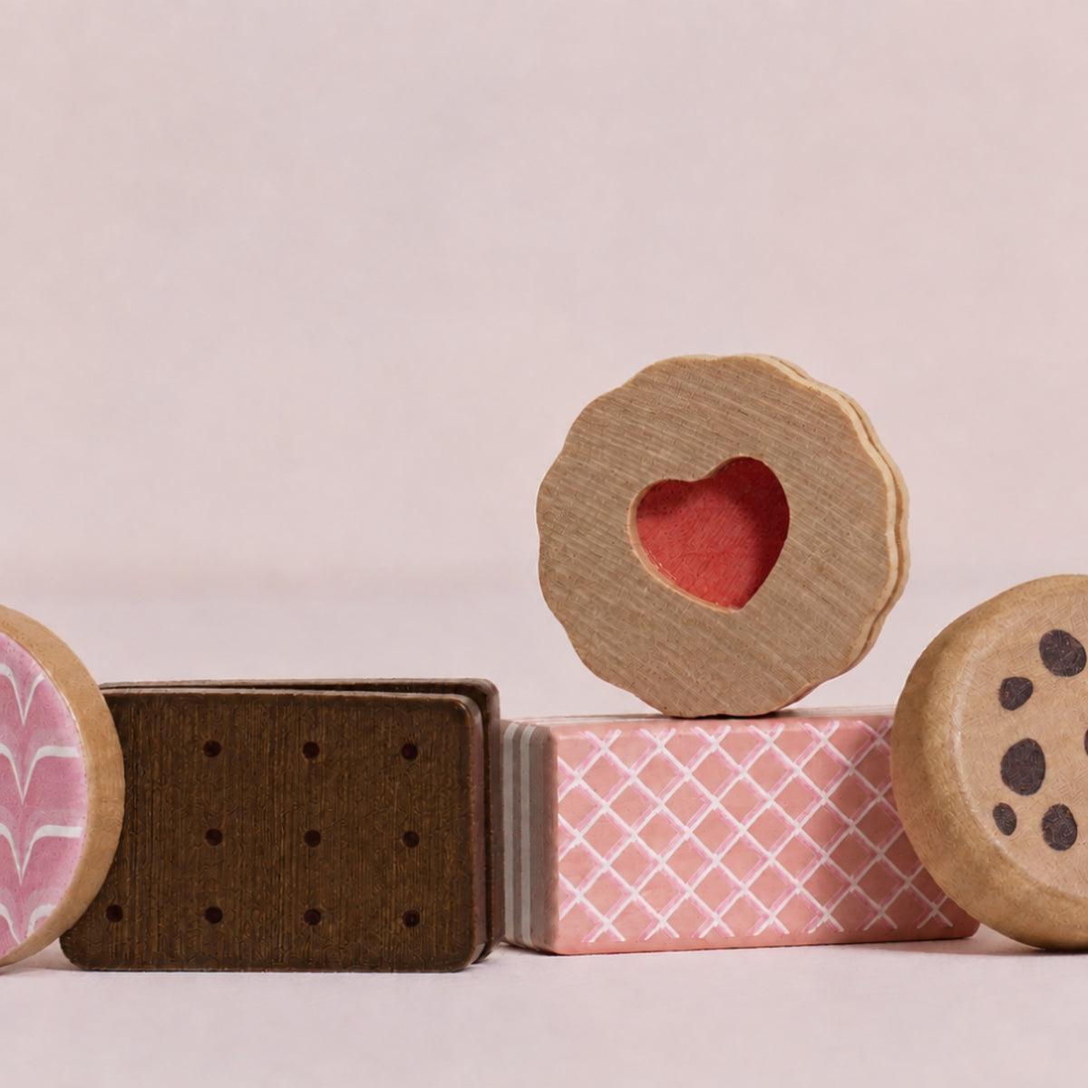 A set of wooden toy sandwich biscuits, including a heart-shaped biscuit on top of a pink wafer, with other toy cookies partially visible on both sides, set against a solid pale pink background.
