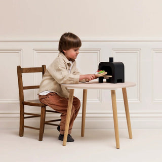A young child sits on a wooden chair at a small round table, playing with a black toy pizza oven and pizza base. The scene is set against a clean, cream-colored paneled wall.