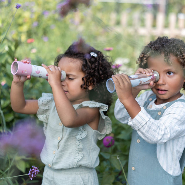 Two young children outdoors in a garden, each holding and looking through a small kaleidoscope. The child on the left wears a pale sage green dress, and the child on the right wears a white button-front shirt with a dusty blue dunagrees.