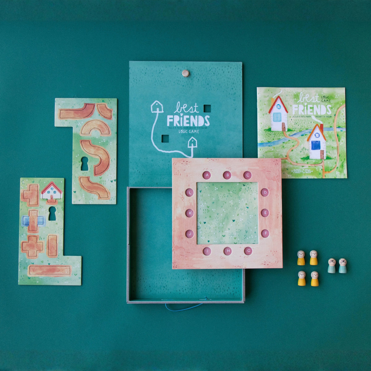 A children's wooden board game set titled 'Best Friends' arranged on a teal background. The set includes a game board with white line art, a pink wooden play square with circular cutouts, several illustrated wooden path and house pieces, and small toy figures.