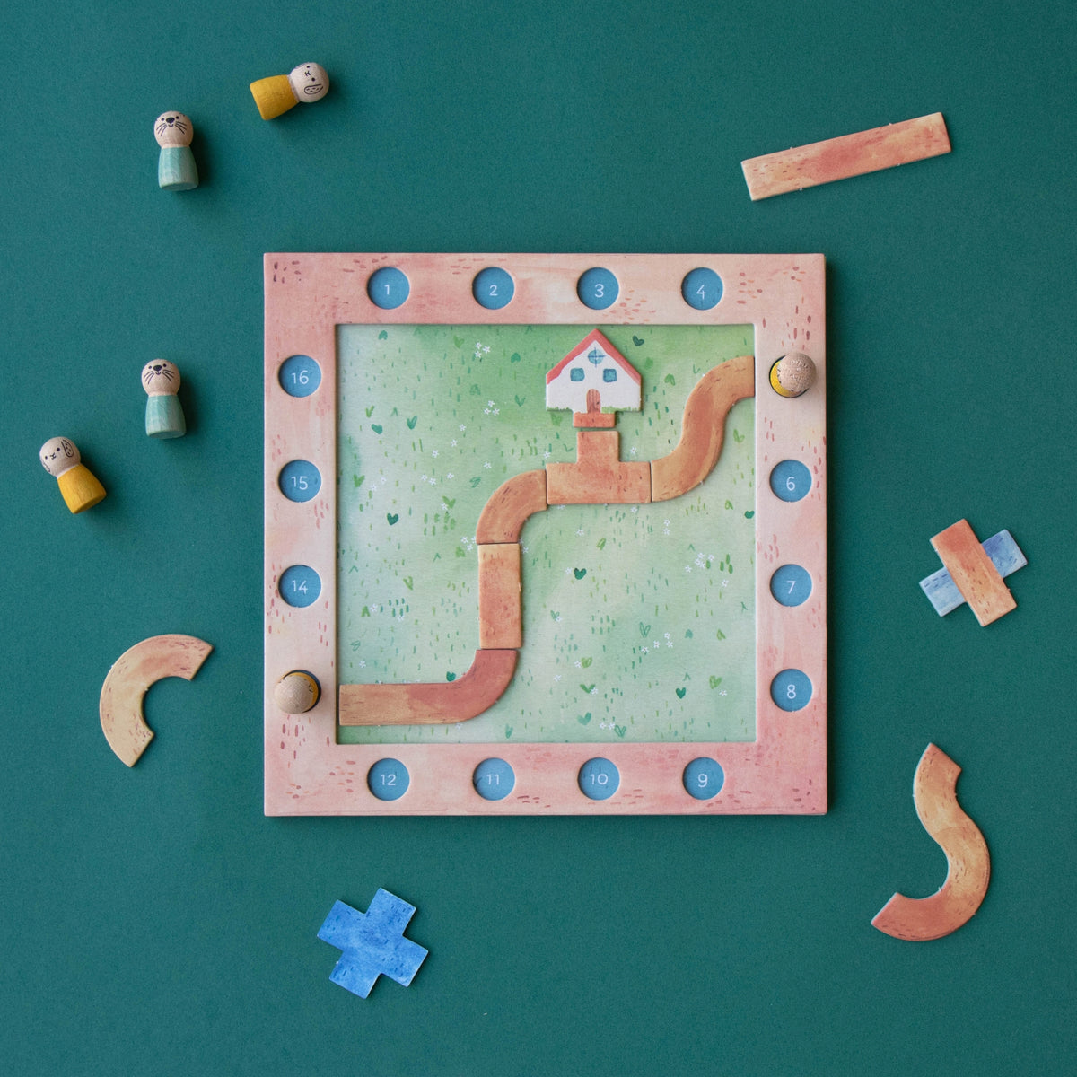 A children's wooden board game set arranged on a dark teal background. The square game board has a pink frame, a light green grassy center, a winding tan path, numbered blue circles around the border, and small house and geometric pieces.