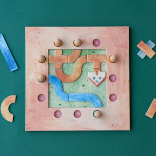 A square wooden puzzle board with a pinkish-beige frame and a colorful geometric maze design, photographed from above on a dark green background with a few loose wooden pieces nearby.