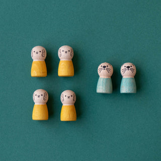 Six small wooden animal-shaped peg figures with painted faces are arranged on a teal background. Four dolls are rabbits with yellow tops and bare wooden heads, and two are otters in light blue tops with bare wooden heads.