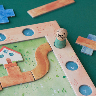 A close-up of a children's wooden board game on a dark green surface. The game features a light wood frame with numbered blue circular spaces, a green play area with a winding tan path, a small house piece, and a small otter-shaped token standing in a hole.