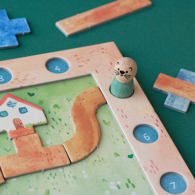 A close-up of a children's wooden board game on a dark green surface. The game features a light wood frame with numbered blue circular spaces, a green play area with a winding tan path, a small house piece, and a small otter-shaped token standing in a hole.