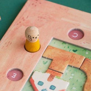A close-up of a wooden children's game board with a small rabbit figure standing in a slot. The board is light peach with painted wooden details, including a green landscape section with matching pieces and pink circular inserts with white letters.