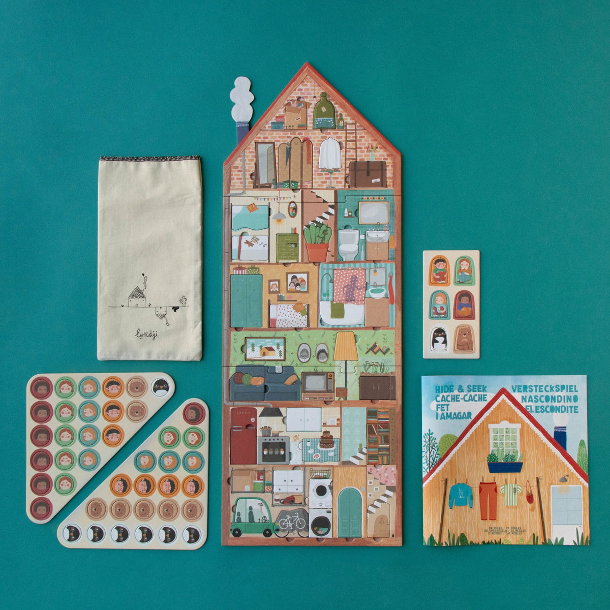 A children's house-themed puzzle arranged on a teal background, including a tall house-shaped board with illustrated rooms, a cream fabric pouch, and threes sheet of round tokens.