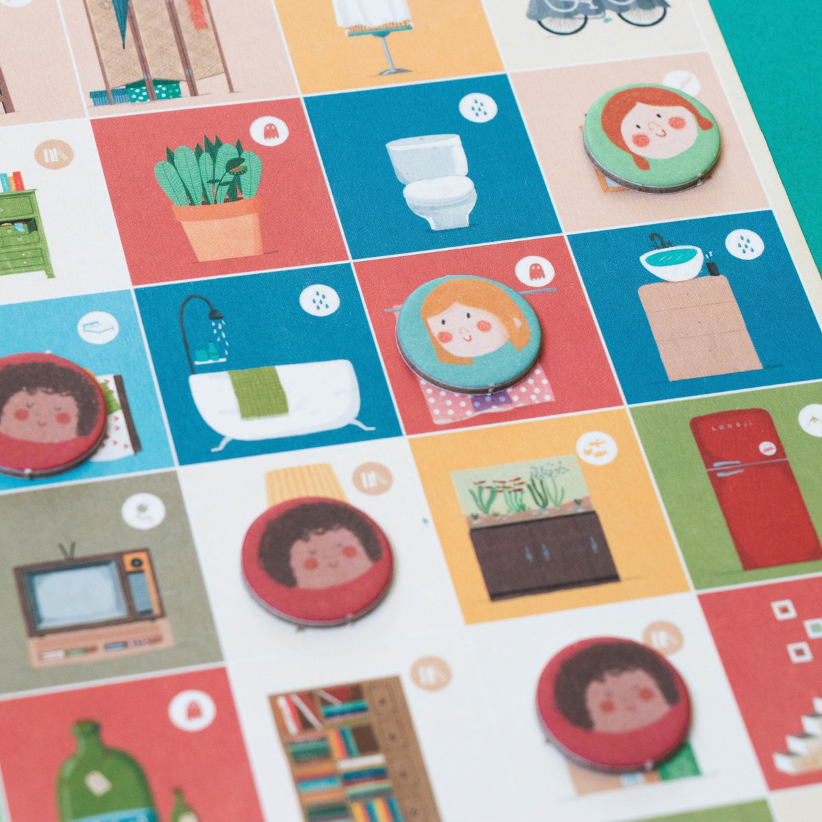 A close-up of a colorful printed board featuring illustrated tiles with objects like a toilet, bathtub, potted plant, TV, books, and round character tokens. 