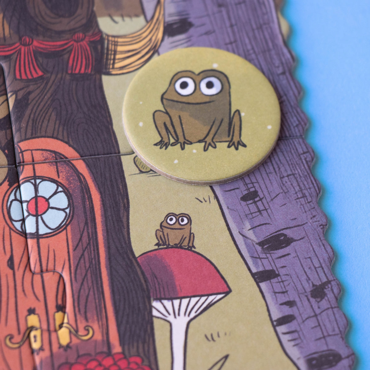 A close-up of a circular token featuring a cartoon frog sitting on a toadstool, placed on a colorful illustrated puzzle with a treehouse and purple birch tree background.