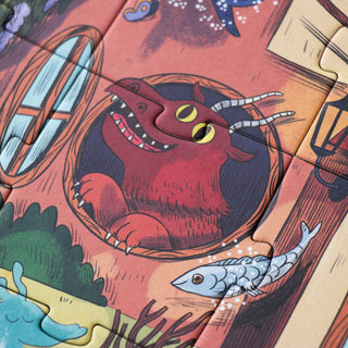 A close-up of a colorful illustrated jigsaw puzzle showing a red monster dragon peeking out of a circular opening, with a fish and fantasy scenery around it.