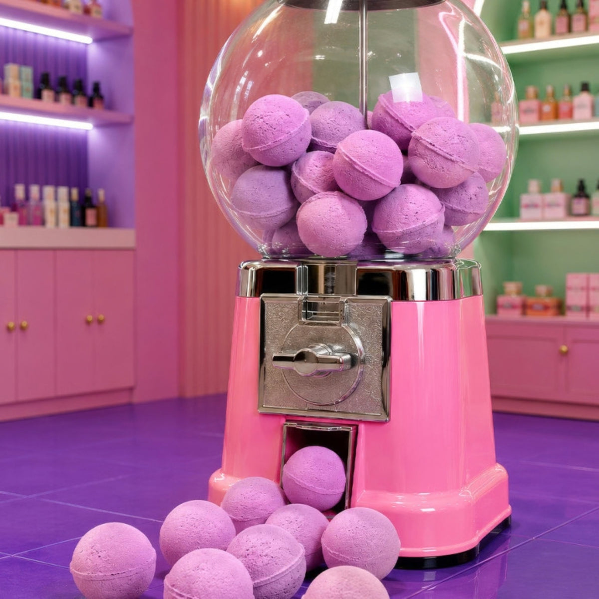 A pink gumball machine filled with purple bath bombs, with several bath bombs spilled onto the floor in front, set in a colorful pink and purple boutique interior.