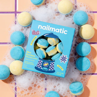 A box of nailmatic mini bath bombs surrounded by blue and pale yellow bath bombs on a peach tiled surface with white bubbly foam.