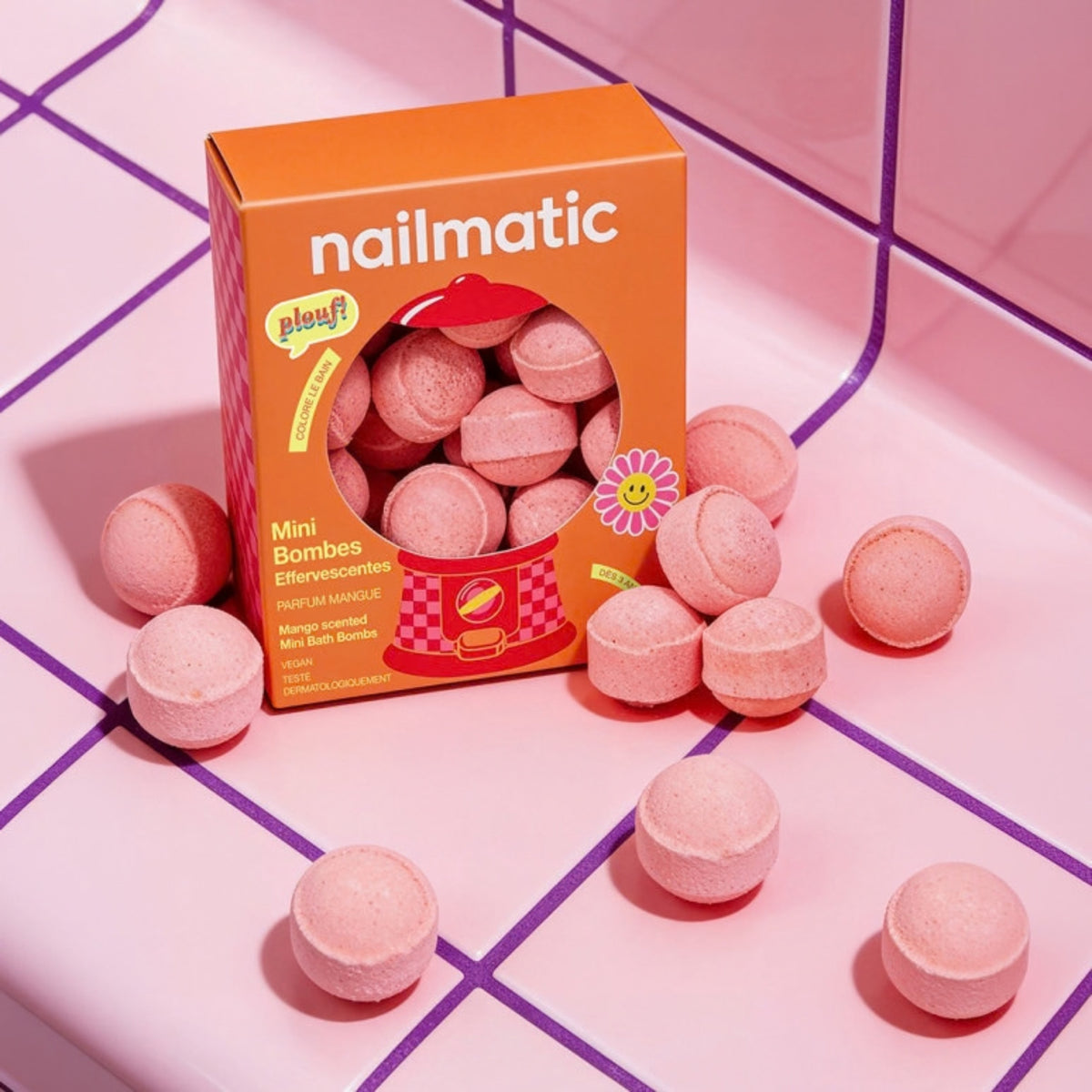 An orange box of Nailmatic mini bombs surrounded by scattered orange round bombs on a pink surface with purple grid lines.