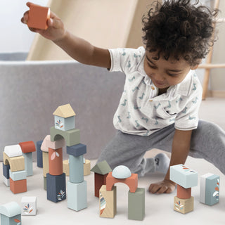 A toddler with curly dark hair sits on a light floor, holding an orange block above a collection of pastel wooden building blocks with arches, towers, and geometric patterns.