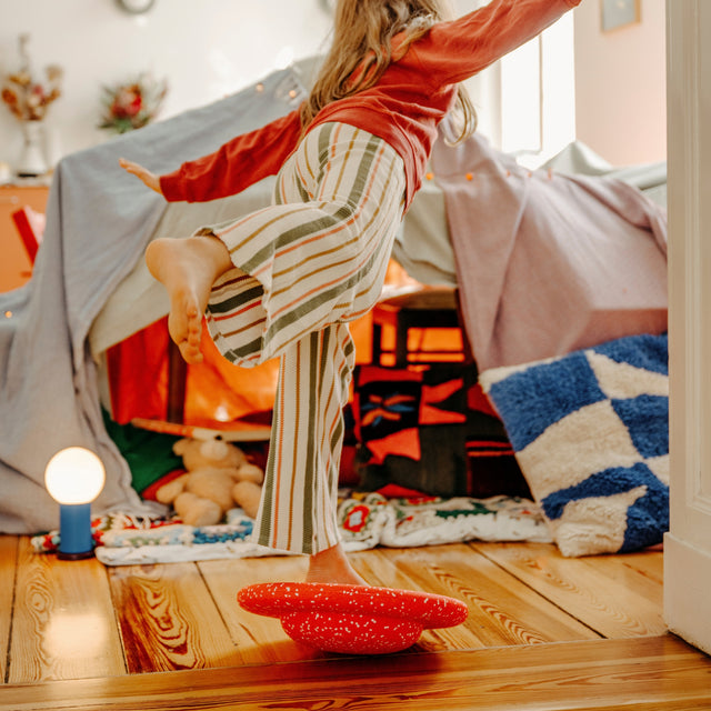 A child wearing a rust-orange long-sleeve top and green, white, and orange striped trousers balances on a red speckled round balance board in a cosy bedroom with a blanket fort and wooden floor.