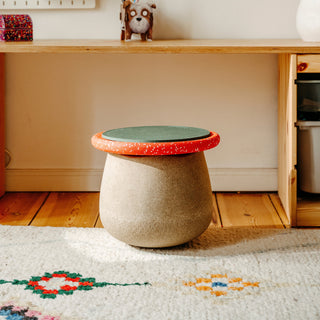 A small round stool with a textured beige body and a lid featuring a green seat pad and red speckled rim, placed on a patterned rug in front of a wooden desk.