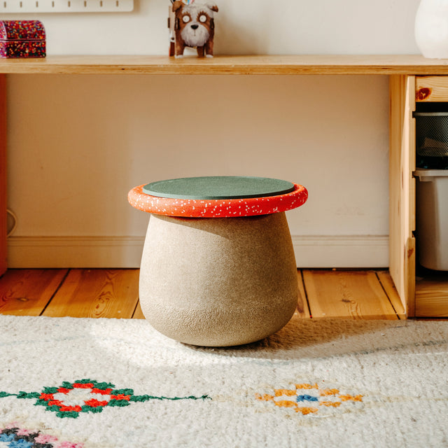 A small round stool with a textured beige body and a lid featuring a green seat pad and red speckled rim, placed on a patterned rug in front of a wooden desk.