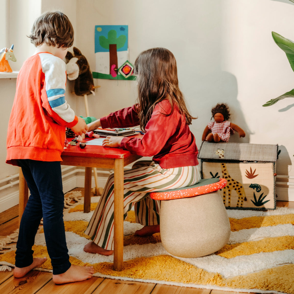 A child's playroom scene with two children interacting at a small wooden table. The child on the left wears an orange and white varsity-style jacket and dark trousers, while the child on the right sits on a round beige toadstool shaped stool with an orange top, wearing a red jacket and striped trousers.