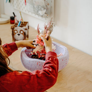 A child wearing a red long-sleeve top arranges a decorative dried floral arrangement in a light purple speckled Original Stapelstein piece.