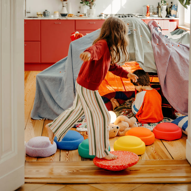 A little girl in a red sweatshirt and striped trousers stands barefoot on a red circular board while another child sits inside a play tent made from blankets, in a bright room with wooden floors and orange cabinets.